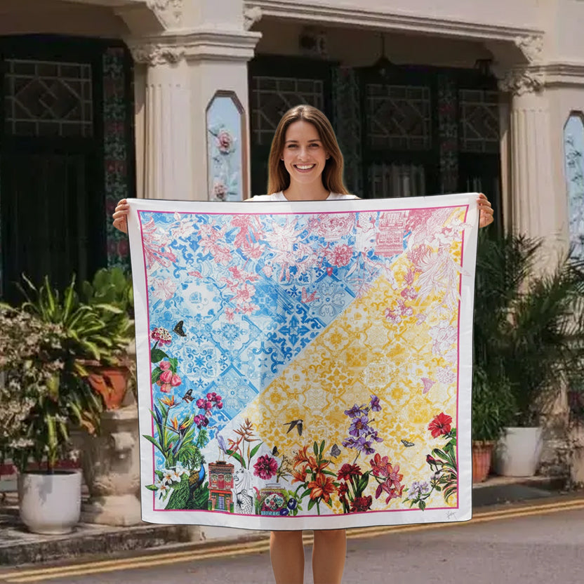 A woman stands outdoors in front of a colonial-style building holding up a large square scarf. The scarf features a colorful, detailed floral and ornamental pattern split diagonally into blue and yellow sections, with a white border and floral motifs along the bottom edge.
