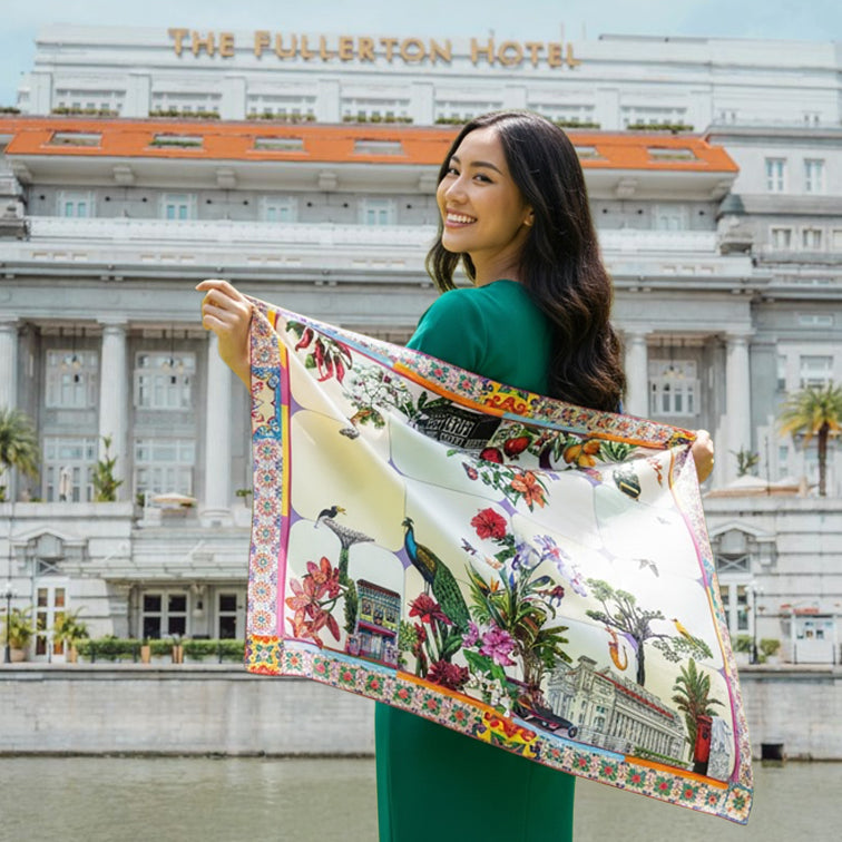A woman in a green dress holds up a large, colorful square silk scarf featuring a tropical garden print with peacocks, flowers, and a classical building, set against the backdrop of The Fullerton Hotel.
